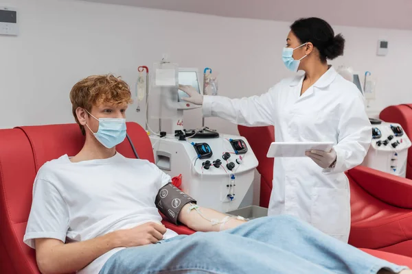 Arzt mit medizinischer Maske hält digitales Tablet in der Hand und bedient Transfusionsmaschine in der Nähe von Rotschopf-Freiwilligen mit Blutdruckmanschette im Labor — Stockfoto
