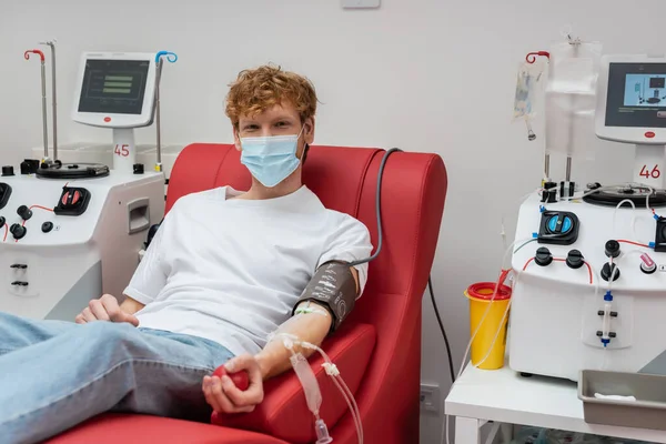 Rothaarige Freiwillige mit medizinischer Maske und Blutdruckmanschette schauen in die Kamera auf einem bequemen Stuhl in der Nähe automatisierter Transfusionsmaschinen im modernen Labor — Stockfoto