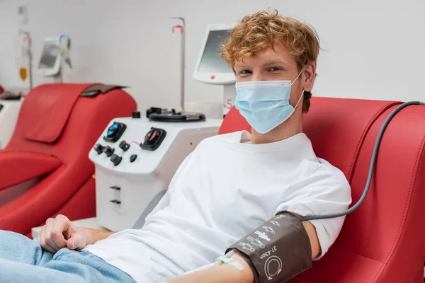 Redhead donor in medical mask and blood pressure cuff looking at camera while sitting on ergonomic chair near automated transfusion machine in hospital — Stock Photo