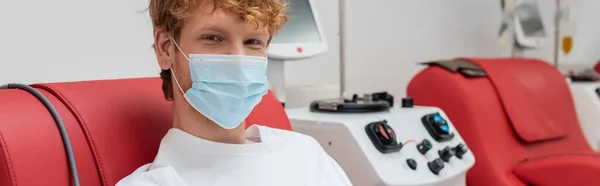 Young and redhead volunteer in medical mask, with positive eyes expression, donating blood near contemporary transfusion machine and looking at camera in laboratory, banner — Stock Photo
