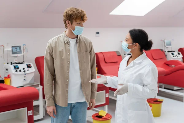 Multiracial doctor in white coat, medical mask and latex gloves talking to redhead volunteer near comfortable chairs and transfusion machines in blood donation center — Stock Photo