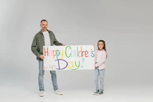 Повна тривалість усміхненого батька і дочки в повсякденному одязі, дивлячись на камеру, тримаючи плакат з щасливим дитячим денним написанням під час святкування разом на сірому фоні — стокове фото