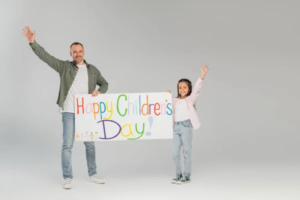 Cheerful father and preteen kid waving hands and holding placard with happy children's day lettering and looking at camera together while standing on grey background — Stock Photo