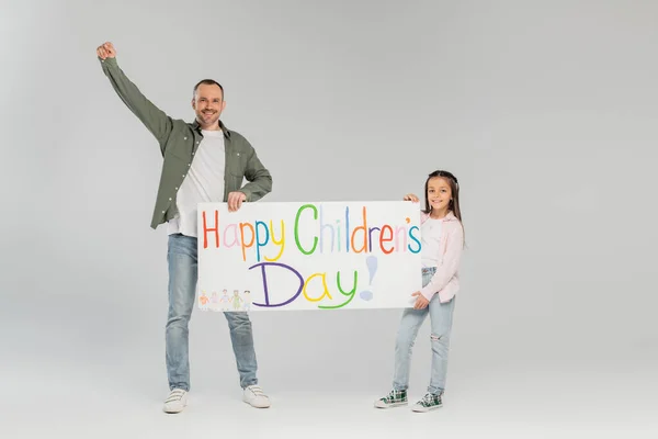 Усміхнена дівчина в повсякденному одязі, що тримає плакат із щасливим дитячим днем, пропускаючи біля батька, показуючи так жест і дивлячись на камеру на сірому фоні — стокове фото