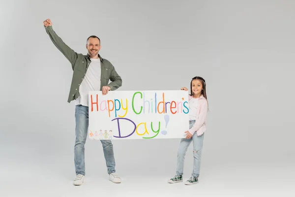 Muchacha preadolescente sonriente con ropa casual sosteniendo pancarta con letras felices del día de los niños cerca de padre mostrando sí gesto y mirando a la cámara en fondo gris - foto de stock