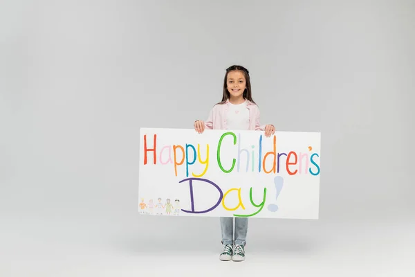 Full length of cheerful preteen girl in casual clothes looking at camera while holding placard with happy children's day lettering while standing on grey background with copy space — Stock Photo