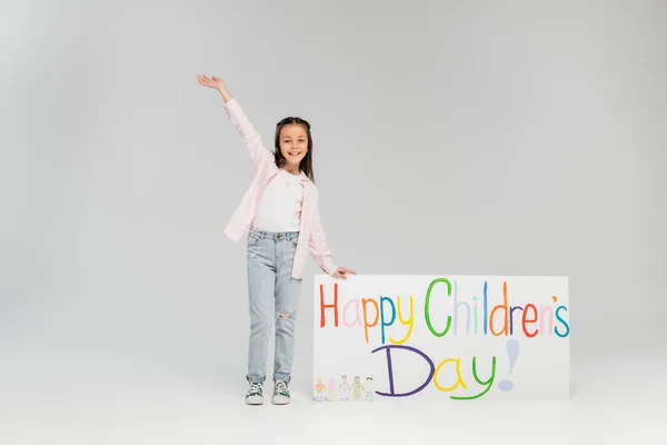 Full length of cheerful preteen girl in casual clothes waving hand and looking at camera near placard with happy children's day lettering and standing on grey background — Stock Photo