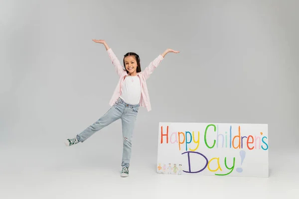 Excited and positive preteen girl in casual clothes having fun and looking at camera near placard with happy children's day lettering during celebration on grey background — Stock Photo