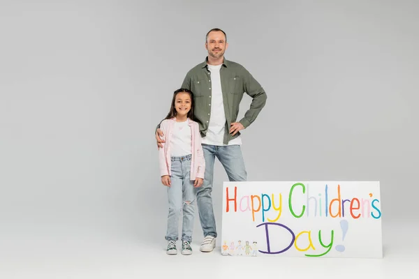 Full length of smiling father in casual clothes hugging preteen daughter and looking at camera near placard with happy children's day lettering on grey background with copy space — Stock Photo