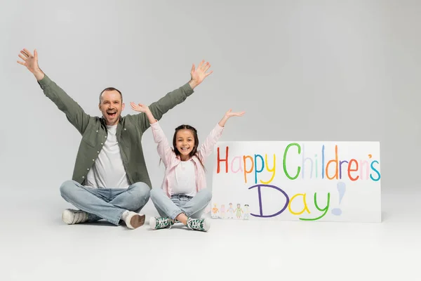 Aufgeregt und fröhlich schauen Mann und Tochter in legerer Kleidung gemeinsam in die Kamera, während sie neben einem Plakat mit fröhlichem Kindertagsaufdruck auf grauem Hintergrund sitzen. — Stockfoto