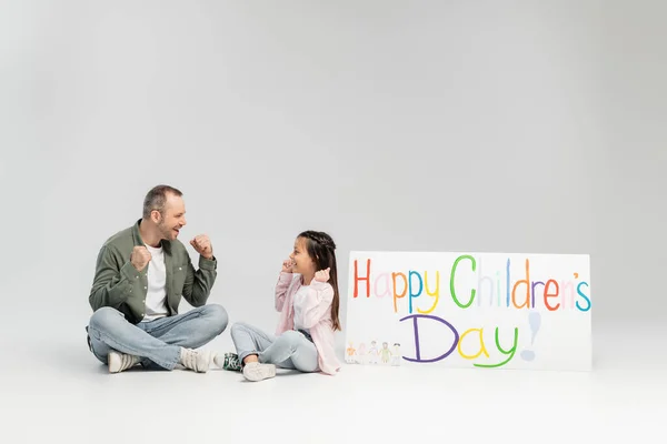 Excited father and daughter in casual clothes looking at each other while sitting near placard with happy children's day lettering during celebration on grey background — Stock Photo
