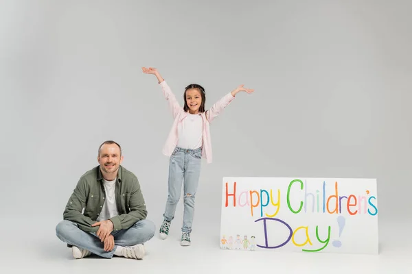 Excited preteen girl in casual clothes looking at camera while standing near father and placard with happy children's day lettering during holiday on grey background — Stock Photo