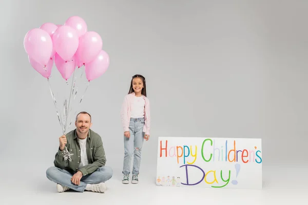 Smiling preteen girl in casual clothes looking at camera near father holding balloons and placard with happy children's day lettering on grey background during celebration — Stock Photo