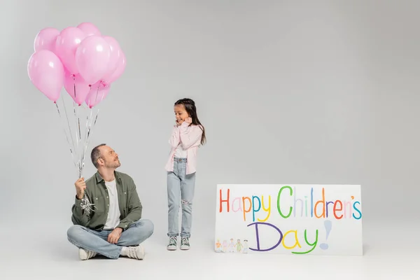 Захоплена дівчинка в повсякденному одязі дивиться на усміхненого батька, який тримає святкові рожеві кульки біля плакату з щасливим дитячим днем, вимовляючи під час святкування на сірому фоні — стокове фото