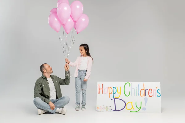 Smiling father giving festive balloons to preteen daughter in casual clothes near placard with happy children's day lettering during event in June on grey background — Stock Photo