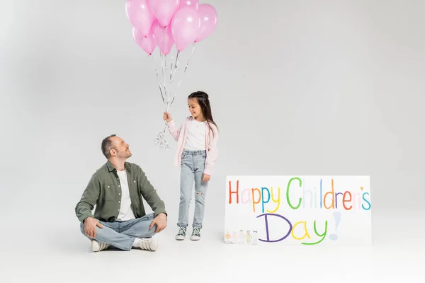 Lächelndes Mädchen mit festlichen Luftballons und Blick auf Vater in legerer Kleidung, der neben Plakat mit fröhlichem Kindertagsaufdruck sitzt, während er im Juni auf grauem Grund feiert — Stockfoto