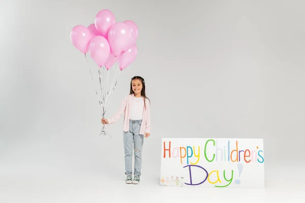 Voller Länge lächelndes Frühchen in lässiger Kleidung mit rosa Luftballons und Blick in die Kamera in der Nähe eines Plakats mit fröhlichem Kindertagsaufdruck während der Feier auf grauem Hintergrund — Stockfoto