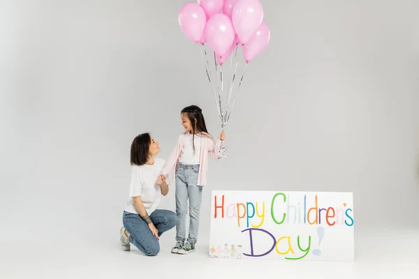 Seitenansicht einer lächelnden Mutter in lässiger Kleidung, die Hand ihrer Tochter mit rosa Luftballons in der Nähe eines Plakats mit fröhlichem Kindertagsaufdruck während der Feier im Juni auf grauem Hintergrund hält — Stockfoto