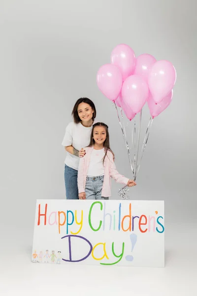 Lächelnde tätowierte Mutter in lässiger Kleidung umarmt ihre Tochter mit rosa Luftballons neben einem Plakat mit fröhlichem Kindertagsaufdruck und feiert im Juni auf grauem Hintergrund — Stockfoto