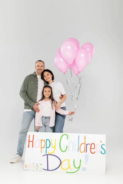 Smiling parents in casual clothes hugging preteen daughter with pink balloons near placard with happy children's day lettering while celebrating together in June on grey background — Stock Photo