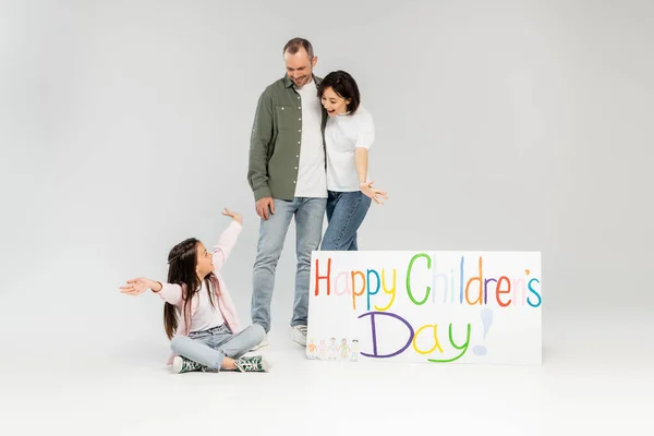 Aufgeregte Eltern schauen bei der Feier im Juni in legerer Kleidung neben einem Plakat mit fröhlichem Kindertagsaufdruck auf grauem Hintergrund auf ihre Tochter. — Stockfoto