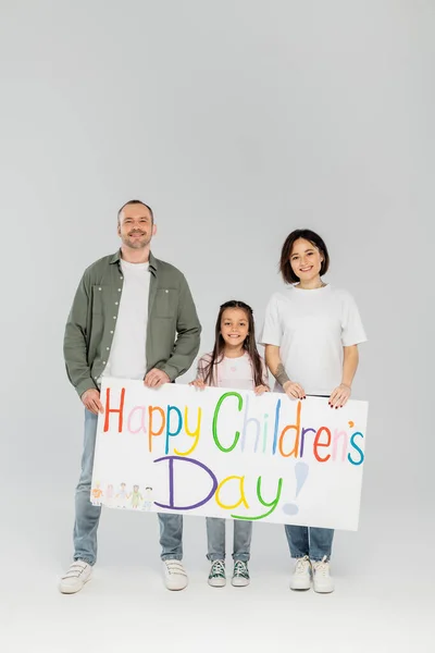 Full length of smiling preteen girl in casual clothes looking at camera while holding placard with happy children's day lettering with parents during event in June on grey background — Stock Photo