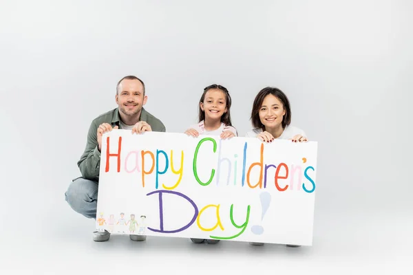 Smiling family with preteen daughter looking at camera while holding placard with happy children's day lettering during celebration together in June on grey background — Stock Photo