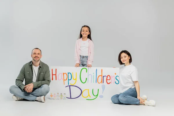 Pleine longueur de pré-adolescent en vêtements décontractés regardant la caméra tout en se tenant près de parents souriants et une pancarte avec le lettrage heureux de la journée des enfants pendant les vacances sur fond gris — Photo de stock