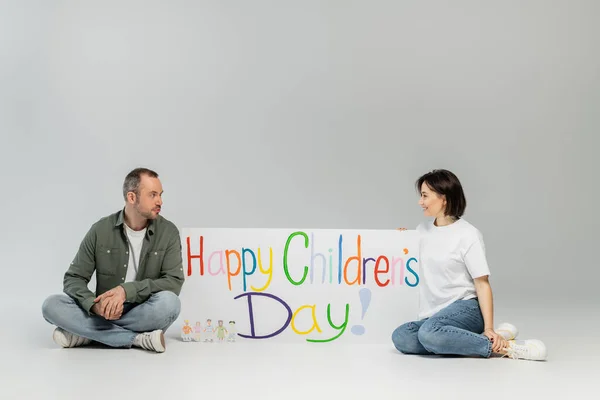 Smiling brunette woman in casual clothes looking at husband while sitting together near placard with happy children's day lettering during celebration on grey background — Stock Photo