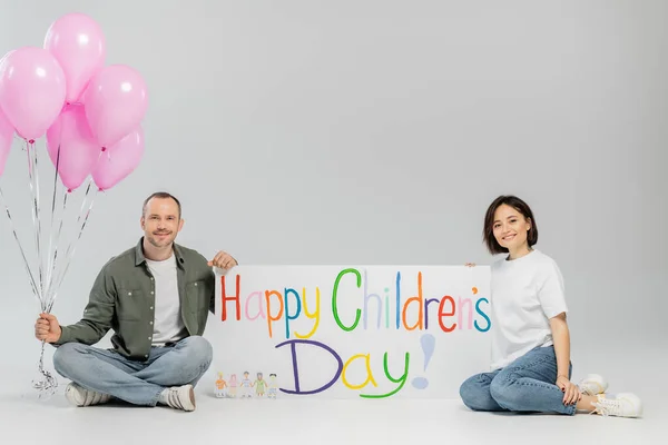 Smiling adult family in casual clothes looking at camera while holding pink festive balloons and placard with happy children's day lettering on grey background with copy space — Stock Photo