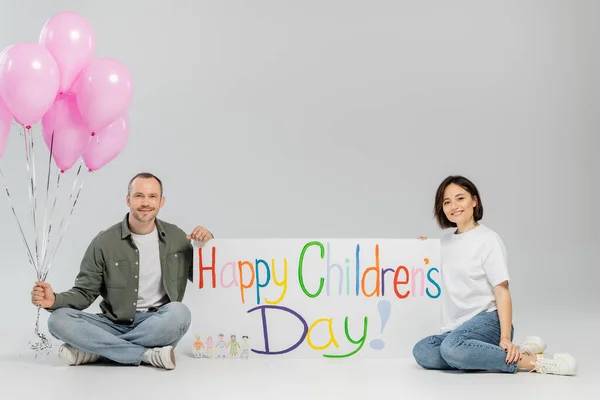 Smiling adult family in casual clothes looking at camera while holding pink festive balloons and placard with happy children's day lettering on grey background with copy space — Stock Photo