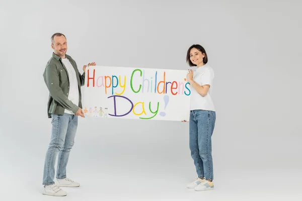 Full length of smiling adult couple in casual clothes looking at camera while holding placard with happy children's day lettering together while standing on grey background — Stock Photo