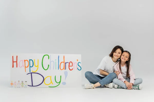 Full length of smiling mother in casual clothes hugging preteen daughter and looking at camera together near placard with happy children's day lettering on grey background — Stock Photo