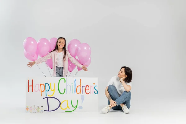 Усміхнена дівчина в повсякденному одязі, що стоїть біля рожевих святкових кульок, плакат з щасливим дитячим днем написання і позитивною мамою, що сидить на сірому фоні під час відпустки — стокове фото