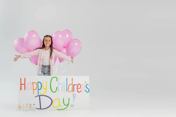 Fille gaie et préadolescente en vêtements décontractés regardant la caméra près de ballons roses festives et pancarte avec lettrage heureux de la journée des enfants tout en célébrant sur fond gris — Photo de stock