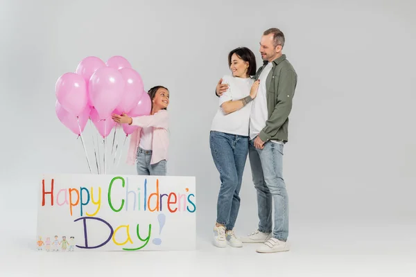 Full length of smiling parents in casual clothes hugging and looking at preteen daughter standing near pink balloons and placard with happy children's day lettering on grey background — Stock Photo