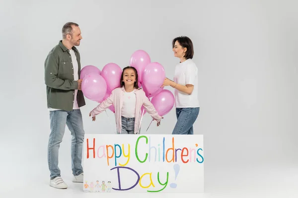 Parents souriants en vêtements décontractés se regardant près de la fille préadolescente, ballons roses et pancarte avec des lettres joyeuses de la journée des enfants sur fond gris — Photo de stock