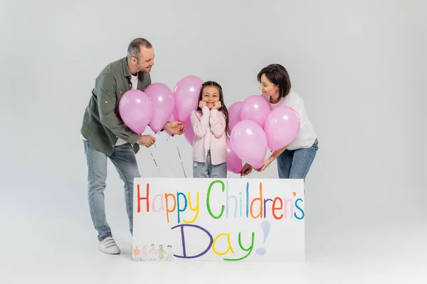 Smiling parents in casual clothes holding pink balloons near cheerful preteen kid and placard with happy children's day lettering during celebration on grey background — Stock Photo