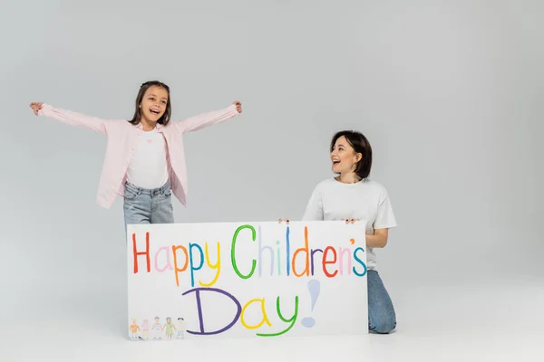 Excited preteen girl in casual clothes looking away while standing near mother and placard with happy children's day lettering during celebration on grey background — Stock Photo
