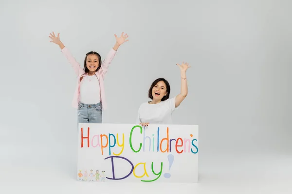 Excited preteen girl in casual clothes waving hands at camera near mother and placard with happy children's day lettering during celebration in June on grey background — Stock Photo