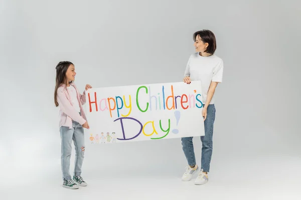 Side view of smiling mother in casual clothes looking at preteen daughter while holding together placard with happy children's day lettering during holiday in June on grey background — Stock Photo