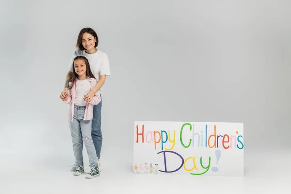 Взрослая женщина в повседневной одежде обнимает веселую дочь-подростка и смотрит в камеру возле плаката со счастливыми детскими буквами во время празднования на сером фоне — стоковое фото