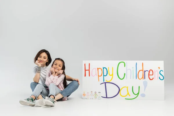 Pleine longueur de mère tatouée souriante regardant la caméra alors qu'elle était assise près de sa fille et une pancarte avec des lettres joyeuses de la journée des enfants pendant la célébration sur fond gris — Photo de stock