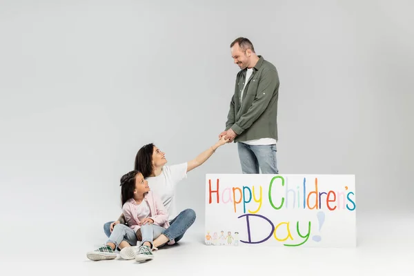 Smiling man holding hand of wife near preteen daughter and placard with happy children's day lettering during kid protection day celebration on grey background — Stock Photo