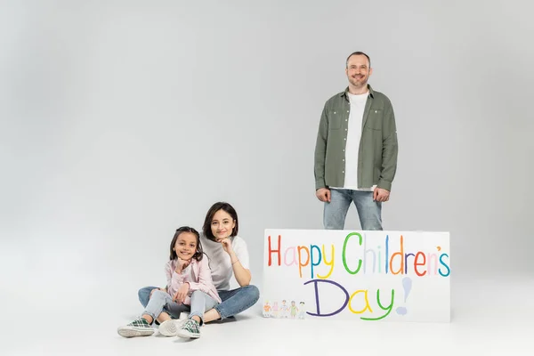 Smiling man looking at camera while standing near wife, preteen daughter and placard with happy children's day lettering during celebration on grey background — Stock Photo