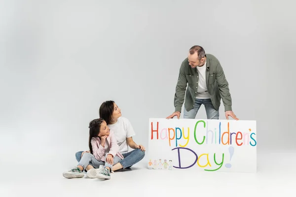 Smiling woman and preteen daughter looking at father standing near placard with happy children's day lettering during celebration in June on grey background with copy space — Stock Photo
