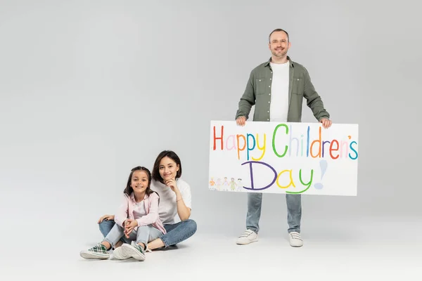 Lächelnder Vater mit Plakat mit fröhlichem Kindertag-Schriftzug in der Nähe von Frau und Tochter, die in die Kamera schauen, während sie im Juni auf grauem Hintergrund sitzen und feiern — Stockfoto