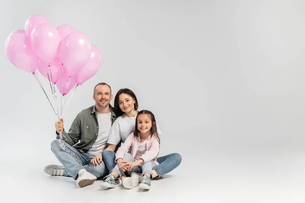 Улыбающиеся родители в повседневной одежде, обнимающие дочку-подростка и держащие праздничные розовые воздушные шары во время празднования международного дня детей на сером фоне — стоковое фото
