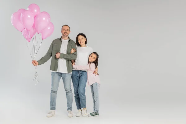 Полная длина веселого человека, держащего праздничные розовые шарики рядом с женой и дочерью-подростком во время празднования Дня защиты детей и глядя на камеру на сером фоне — стоковое фото