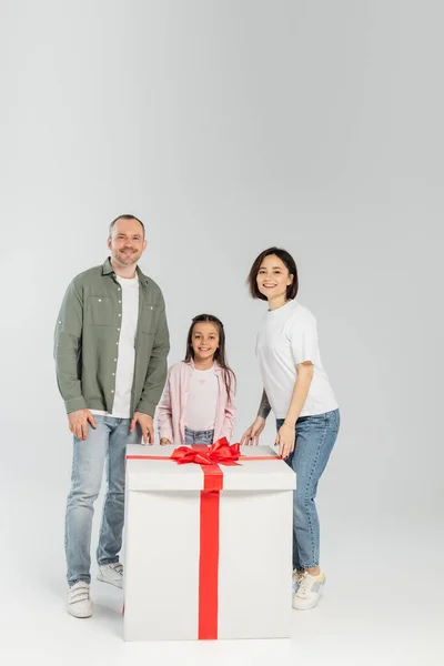 Полная длина улыбающихся родителей и дочери-подростка, смотрящих в камеру рядом с большой подарочной коробкой во время празднования Дня защиты детей на сером фоне — стоковое фото